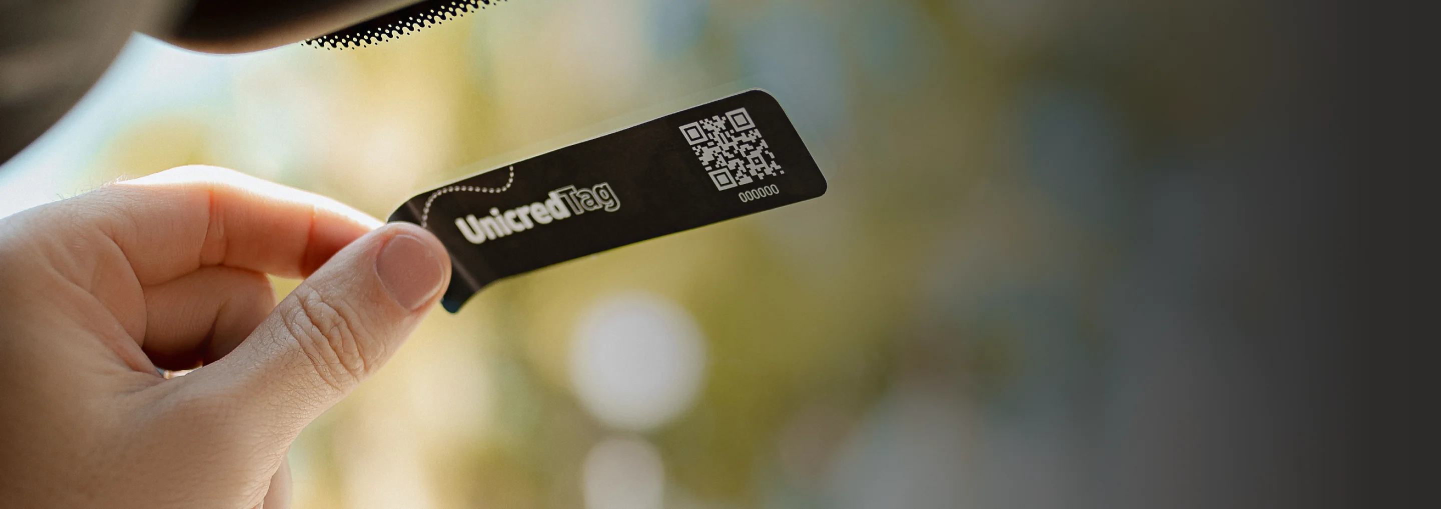 Mão posicionando a Unicred Tag no para-brisa do carro, com fundo externo desfocado.